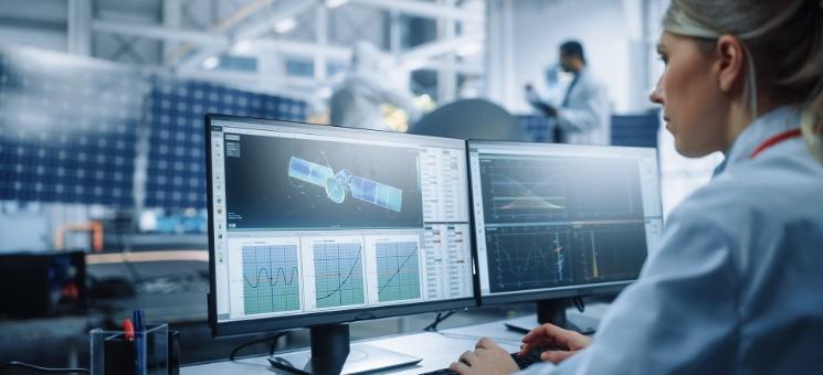 Engineer analyzing satellite data and graphs on dual monitors in a workspace with solar equipment.