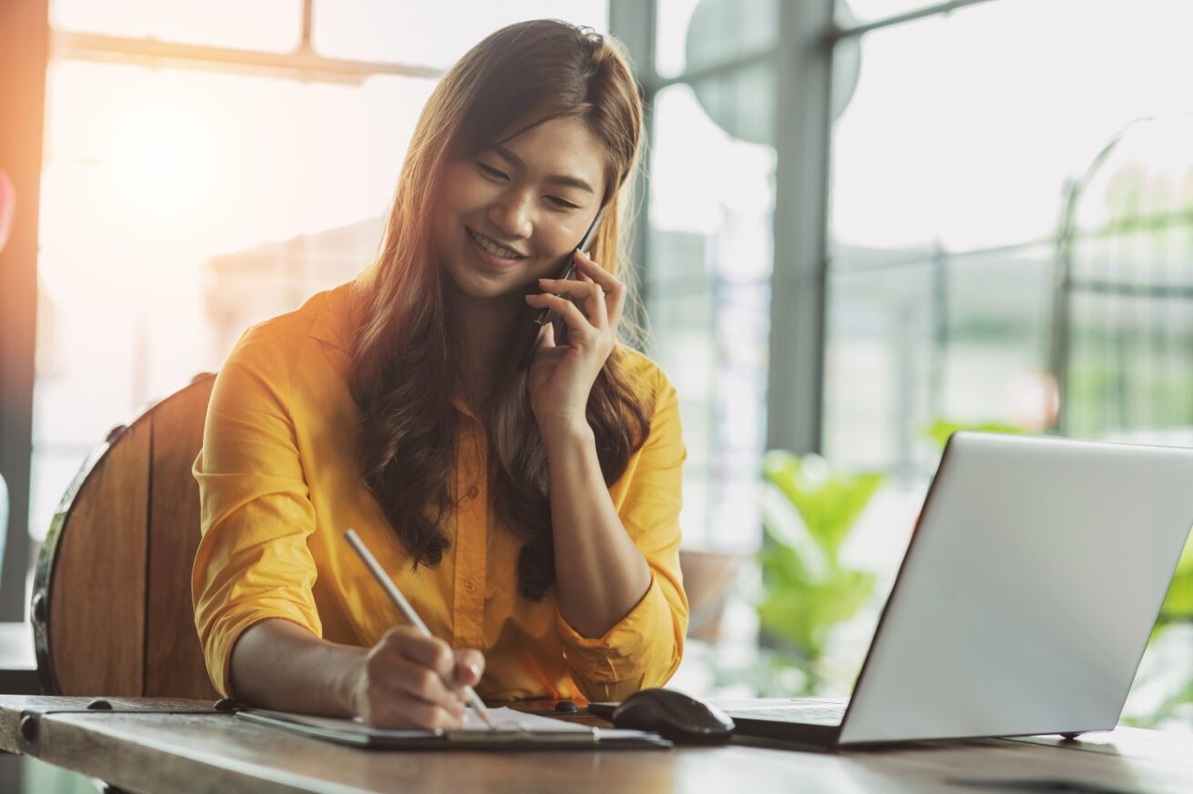 Business woman answering the phone while being sitting on a desk with a computer in front of her