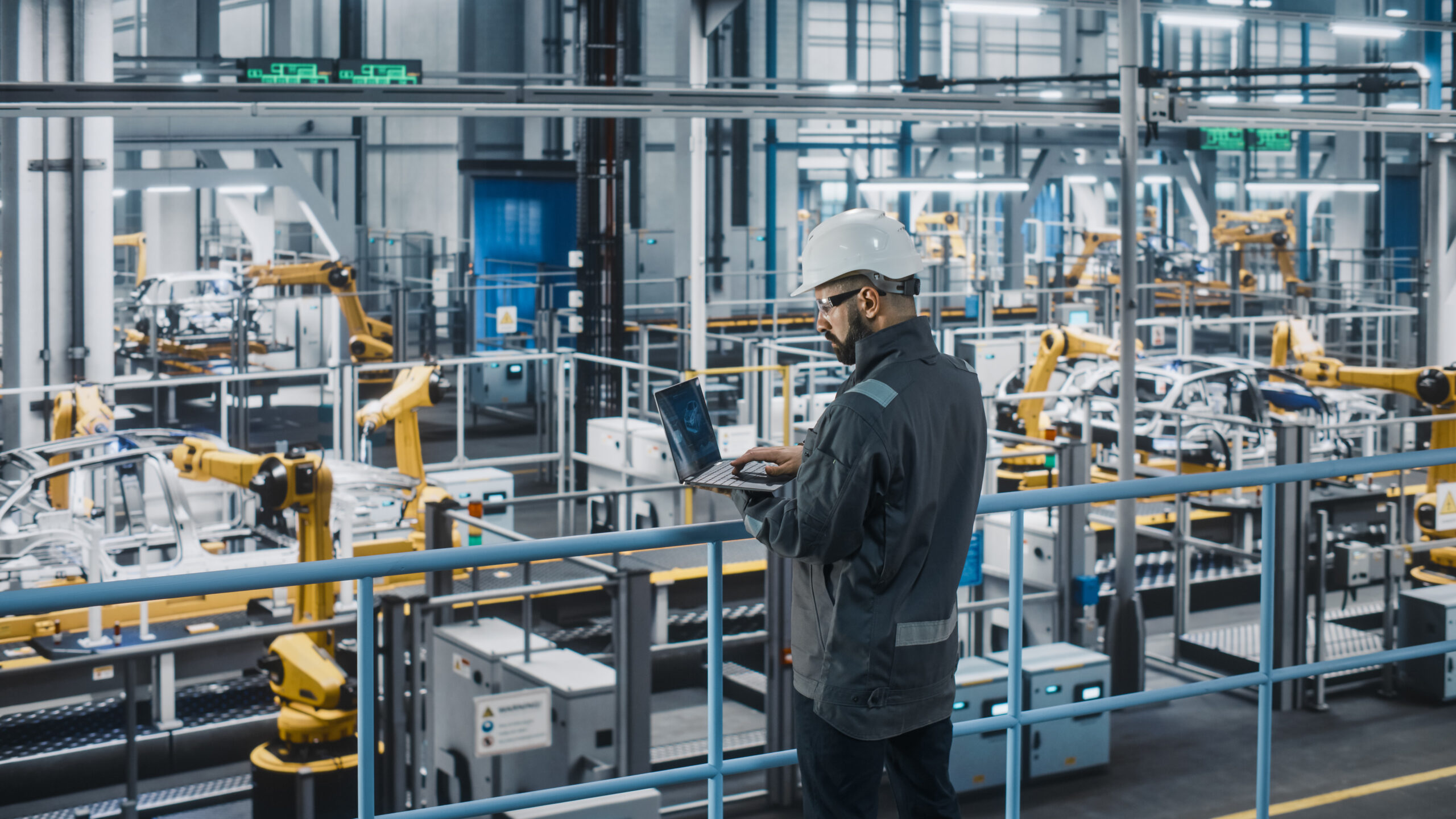 Engineer with laptop on platform overlooking automated factory floor with robotic arms and machinery.