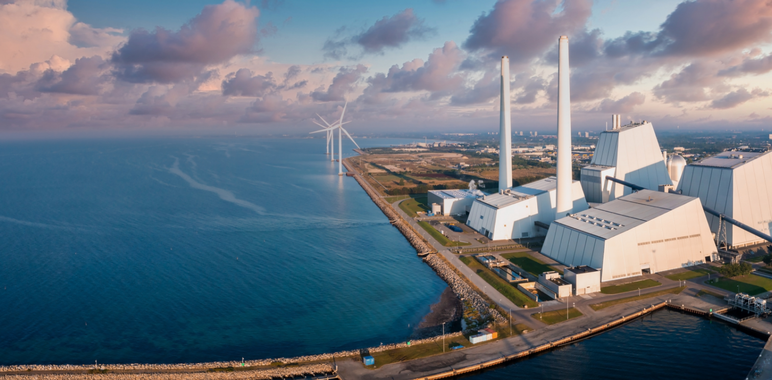 Industrial waterfront with white buildings, chimneys, and wind turbines under partly cloudy sky.
