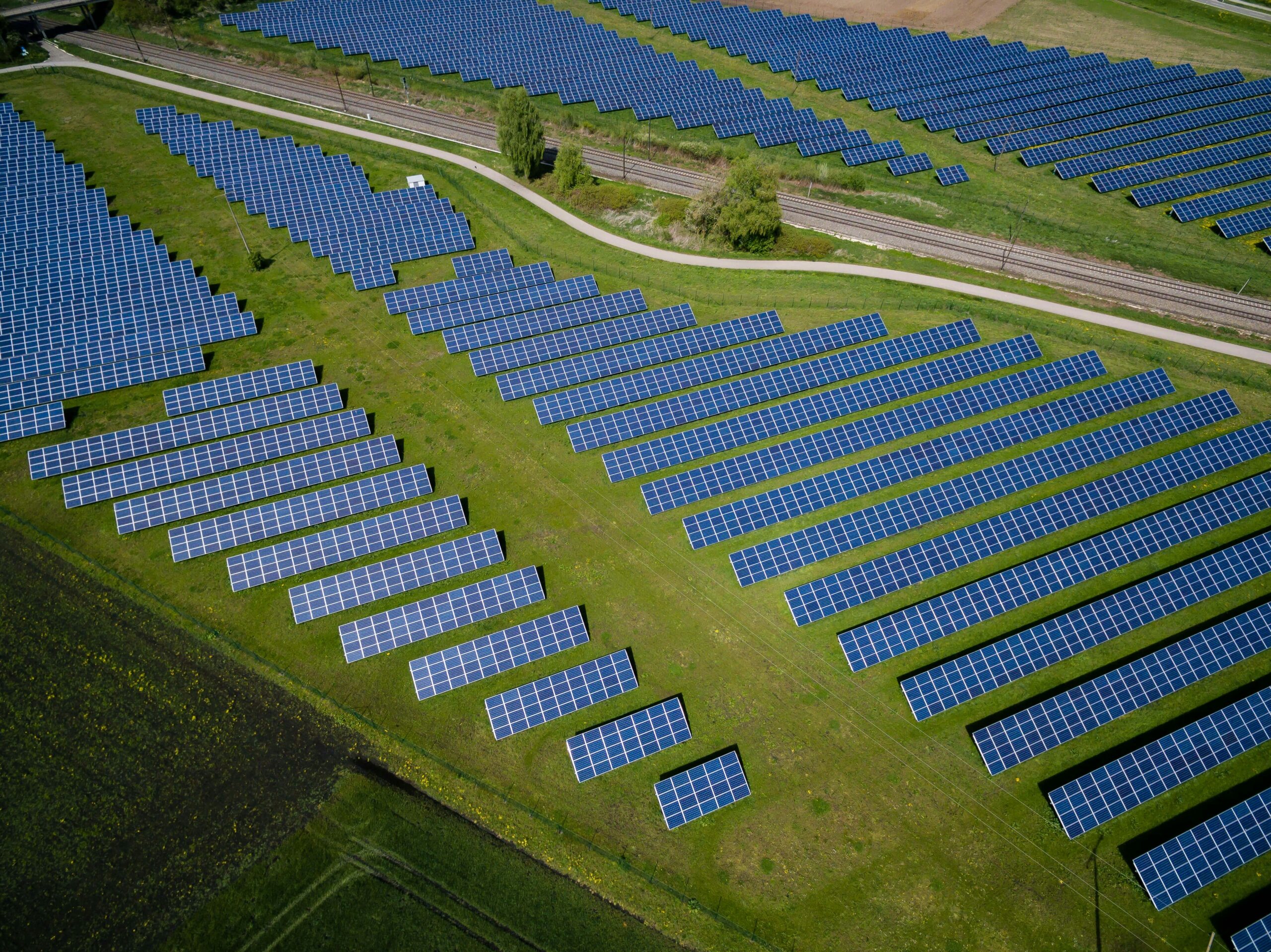 Aerial view of solar farm with blue panels, grassy fields, pathways, and railway track.