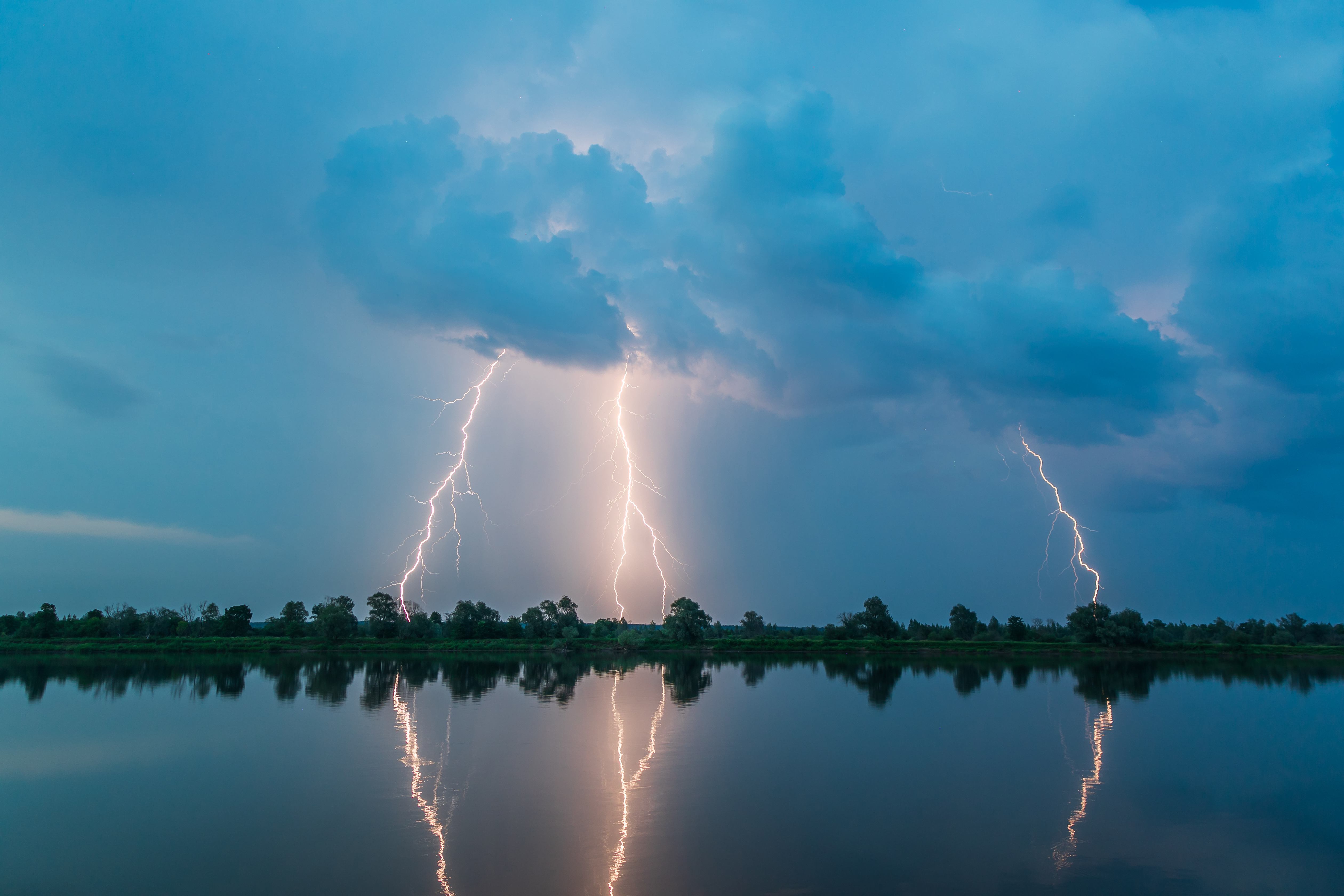 Multiple lightning bolts striking over calm water under dark storm clouds, with reflections on the surface.