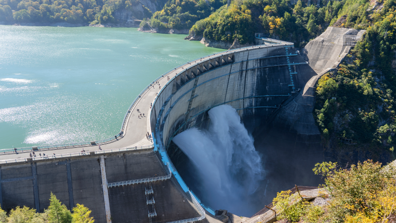Curved dam releasing water in a forested mountain setting with visitors walking along the top.